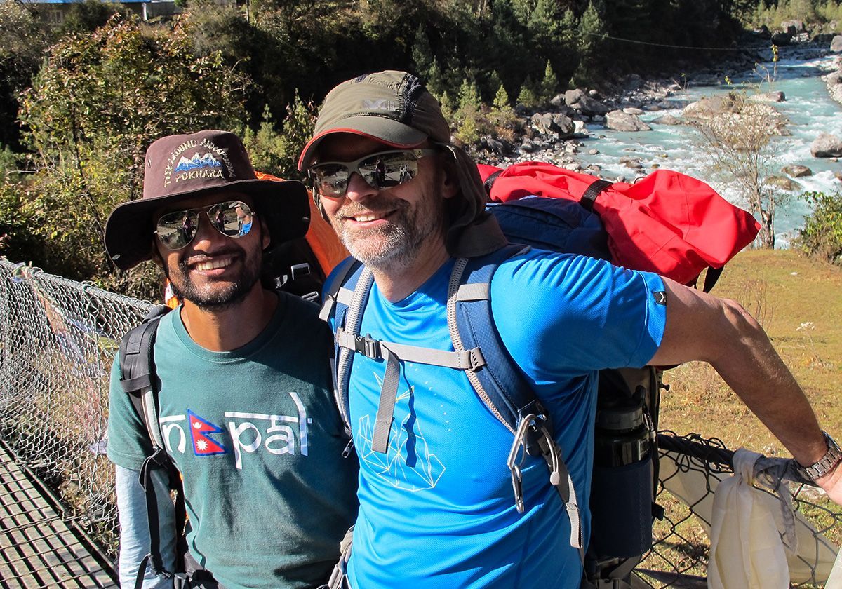 Two trekkers, one in a blue shirt and the other in a Nepal cap, posing happily on a scenic mountain bridge.