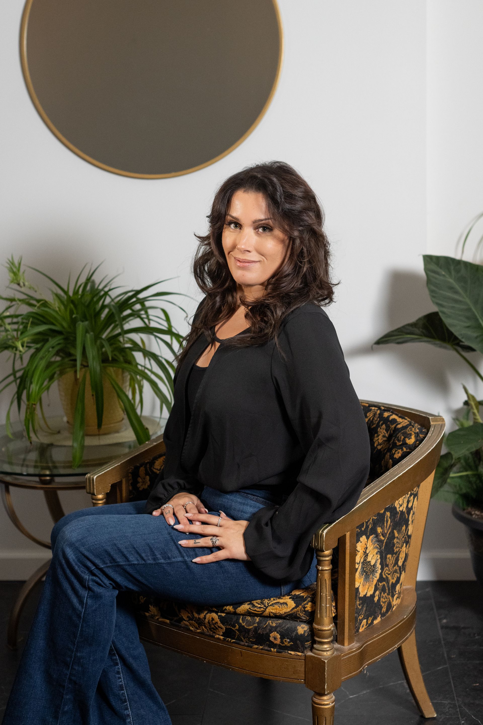 Woman in black top and jeans sits in ornate chair, smiling.