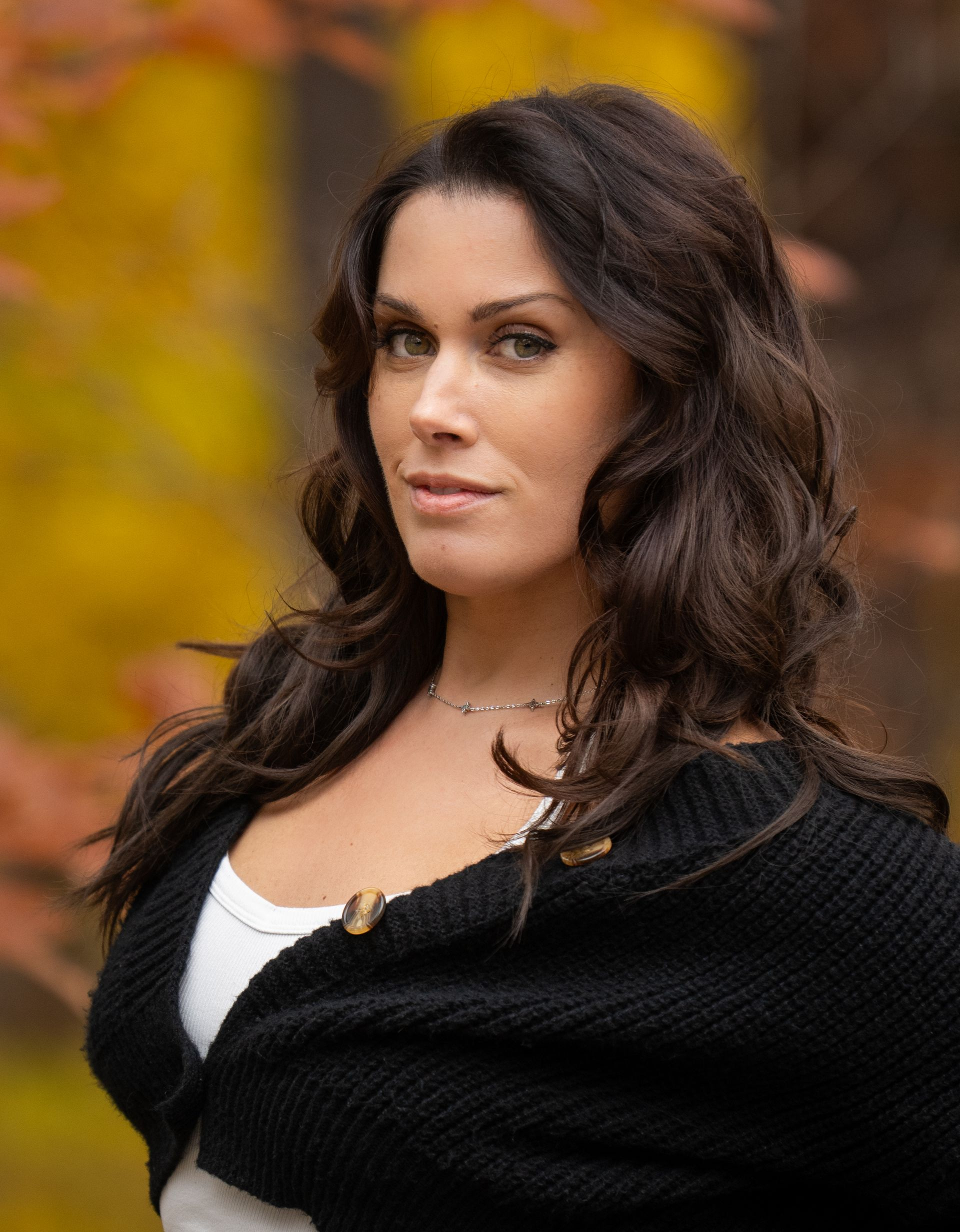 Woman with dark hair in a black sweater, looking at the camera outdoors with fall foliage in the background.