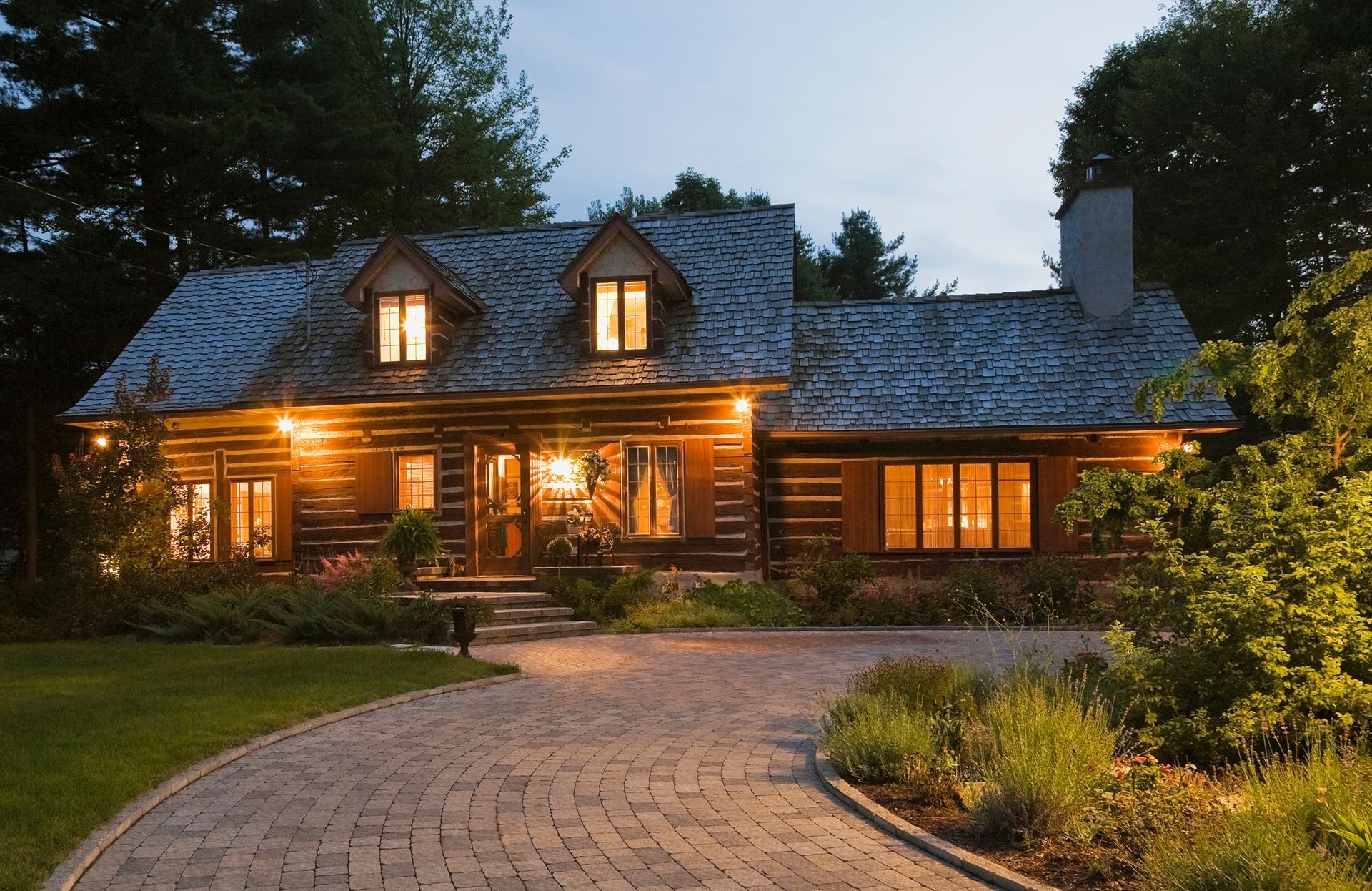 Log cabin at dusk with lights on, cobblestone driveway, surrounded by greenery.