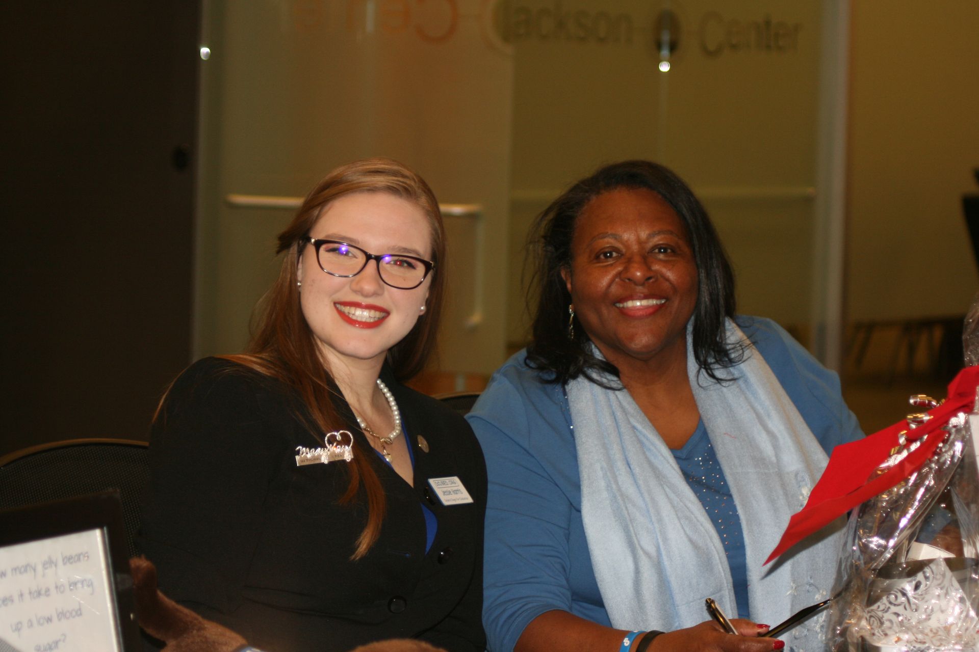 Two smiling people pose for a photo indoors, one wearing a black blazer and glasses and the other a blue top and scarf.