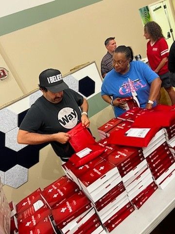 Two people stand at a table, packing and organizing stacks of red mailers and white boxes in an indoor setting.