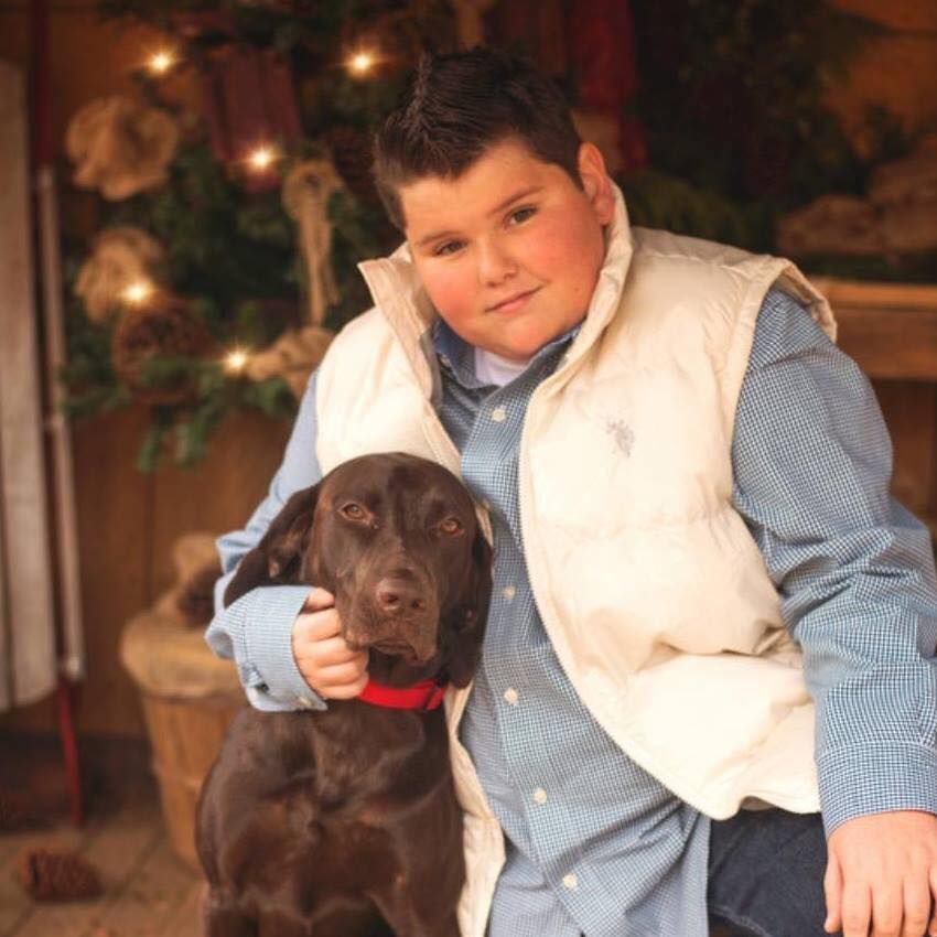 A person in a light vest and blue shirt poses with a brown dog wearing a red collar, set against a festive backdrop.