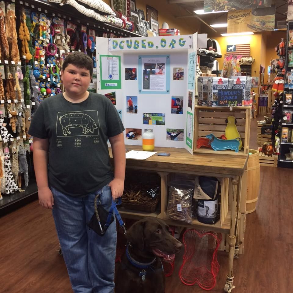 A person stands with a brown dog in a pet supply store beside a display board for DCUBED.ORG.