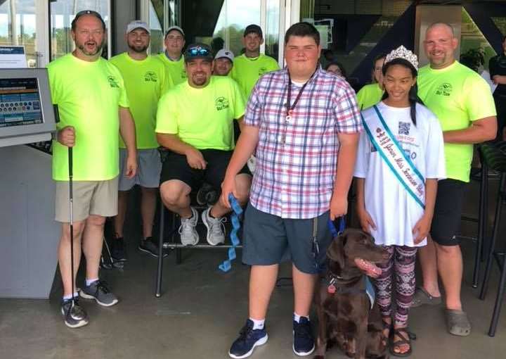 A group of people in neon yellow shirts and one person wearing a sash and crown stand with a brown dog in an indoor space.