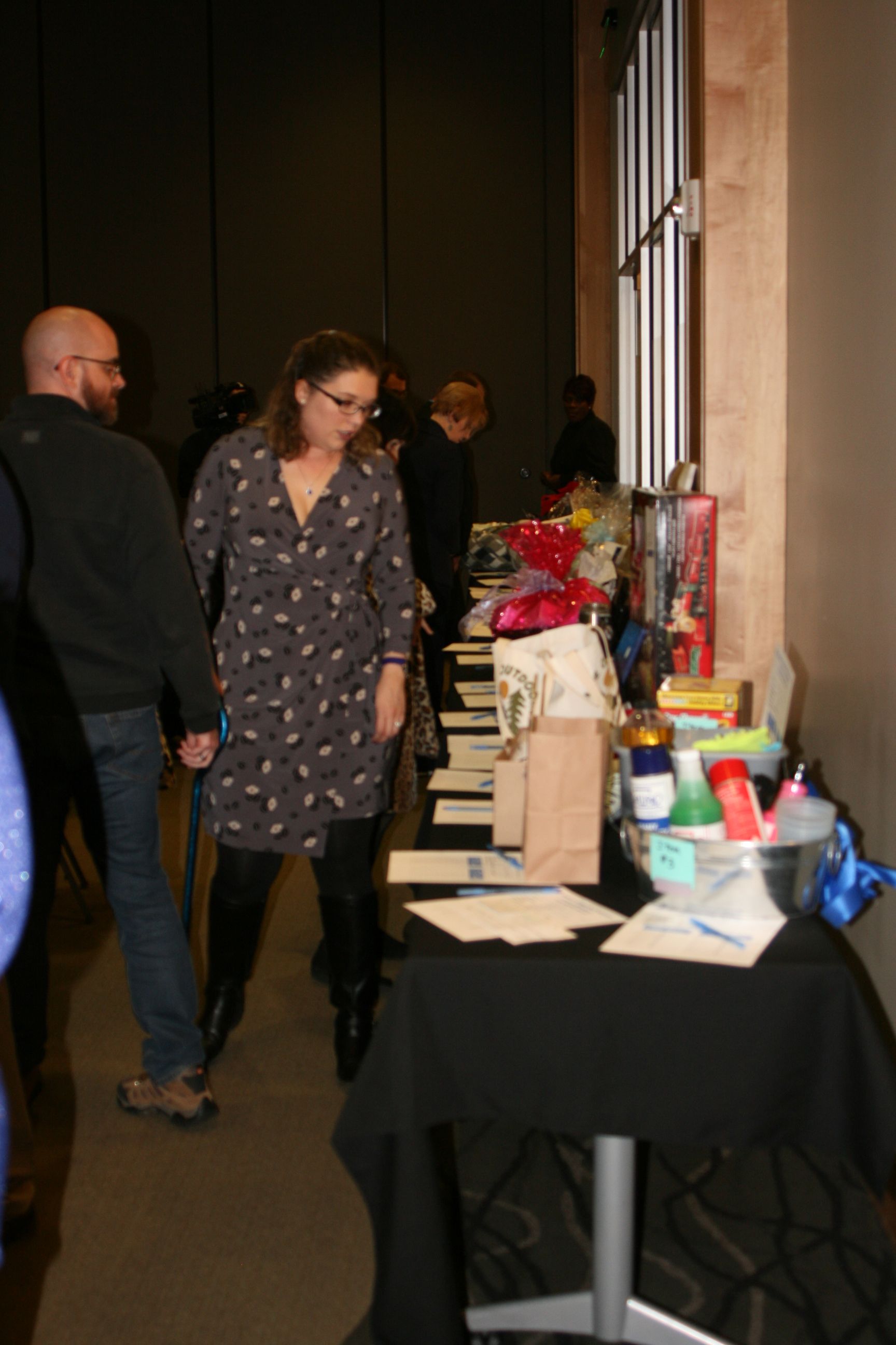 People browsing a silent auction table with gift baskets and items on a black-covered table in an indoor event space.