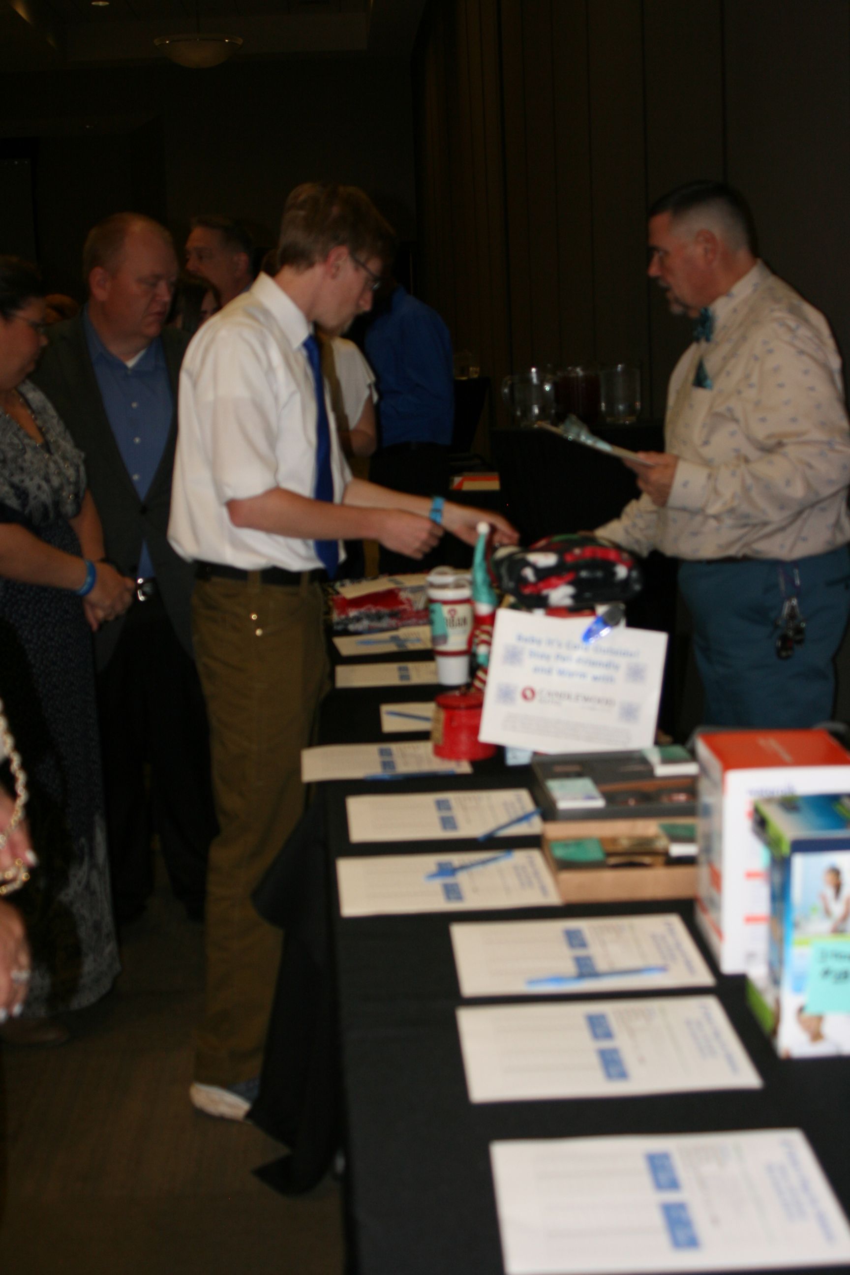 An event attendee speaks with a booth representative across a black table covered with brochures and marketing materials.
