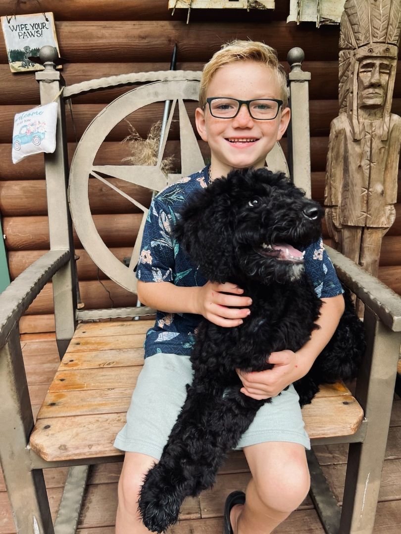 A child with glasses smiles while sitting in a large wooden chair, holding a fluffy black dog on their lap.
