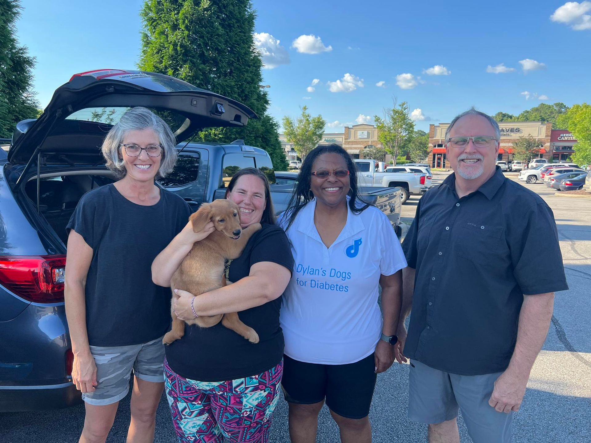 Four people stand outside by an open car trunk; one holds a small, light-brown puppy. They are smiling in a parking lot.