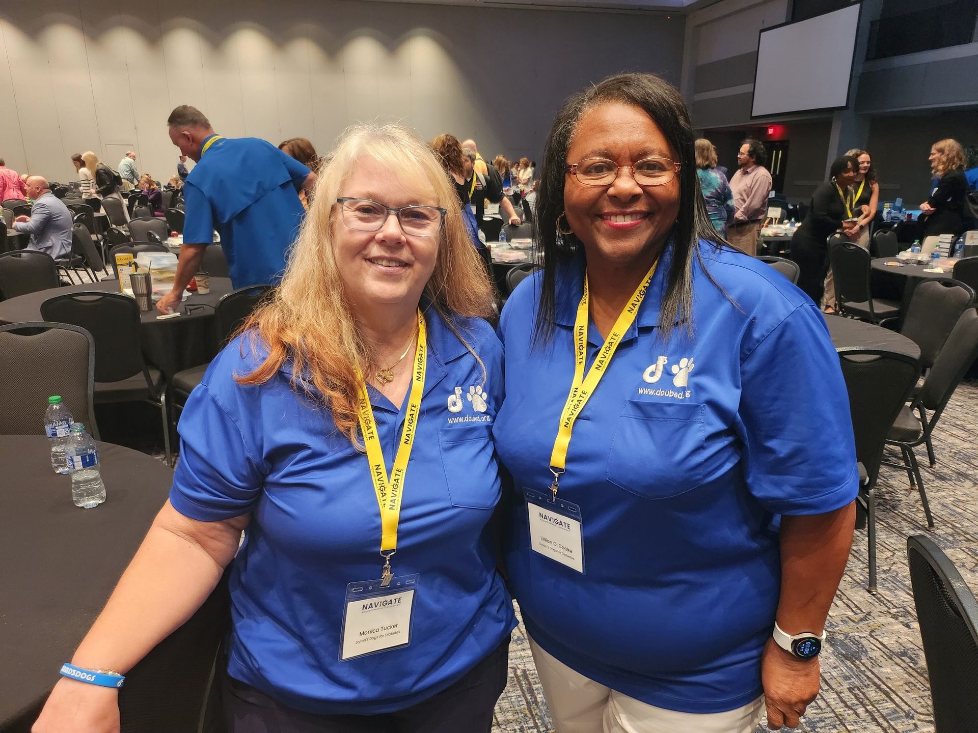 Two people in matching blue polo shirts and yellow lanyards smile for a photo at a large indoor conference.