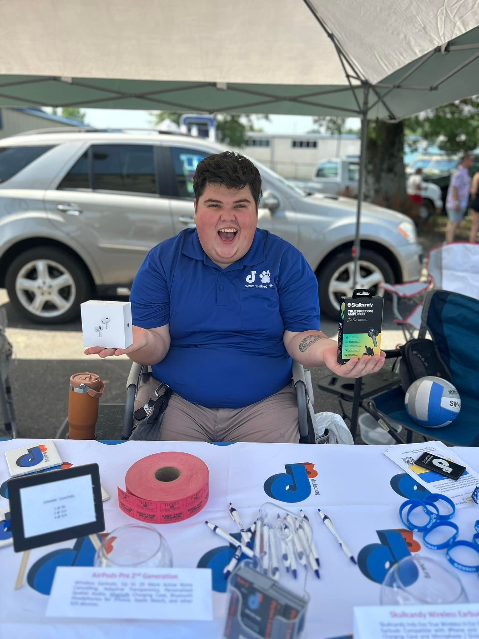 A smiling person in a blue polo shirt sits in a wheelchair behind a promotional table, holding two small electronic boxes.