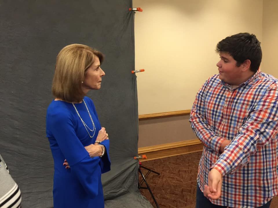 A woman in a blue dress and a person in a plaid shirt stand facing each other in front of a gray backdrop.