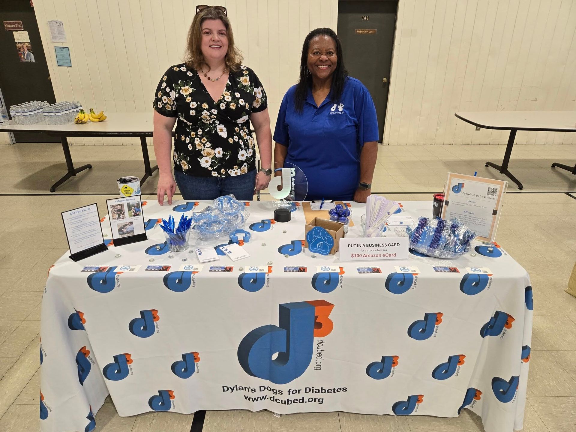 Two people stand behind a table draped in white fabric with blue and orange logos, set up for a community event.