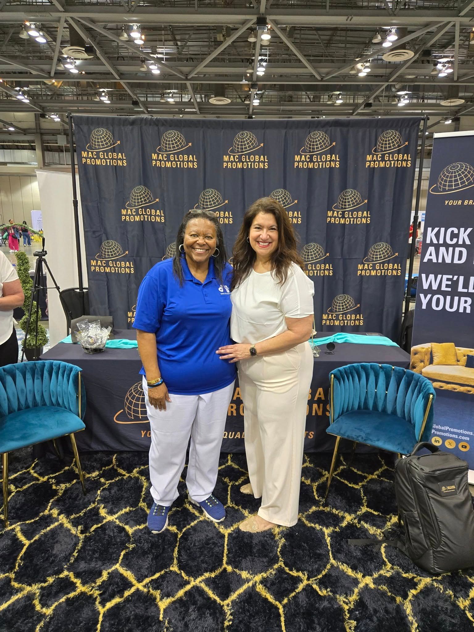 Two people smiling and standing in front of a branded trade show booth with blue velvet chairs and a black patterned rug.