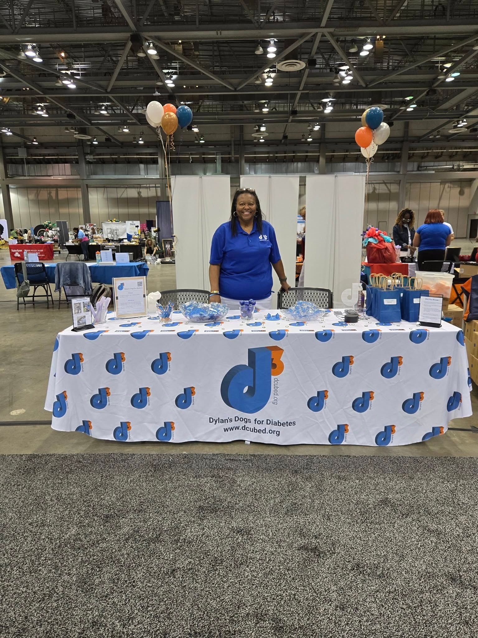 A person stands behind a trade show table with a branded blue logo tablecloth, balloons, and flyers in a large venue.
