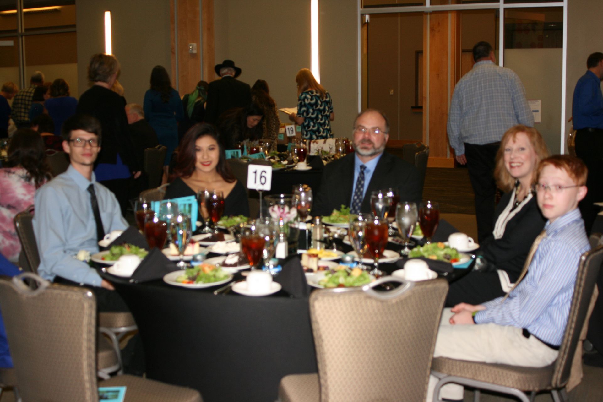 Five people seated at a round dining table in a conference room setting, with a number 16 sign in the center.