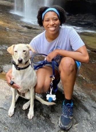 A smiling person crouches by a waterfall, holding a leash attached to a light-colored dog sitting on the wet rocks.