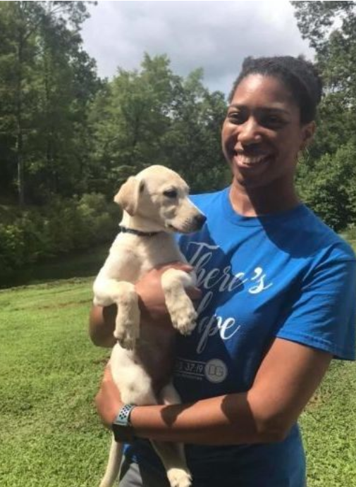 A smiling person holding a light-colored puppy in a green, wooded outdoor setting.
