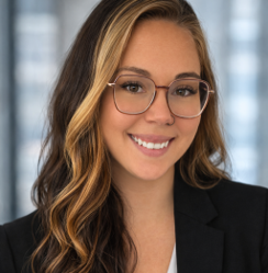 Headshot of a smiling person wearing glasses and a dark blazer against a blurred office background.