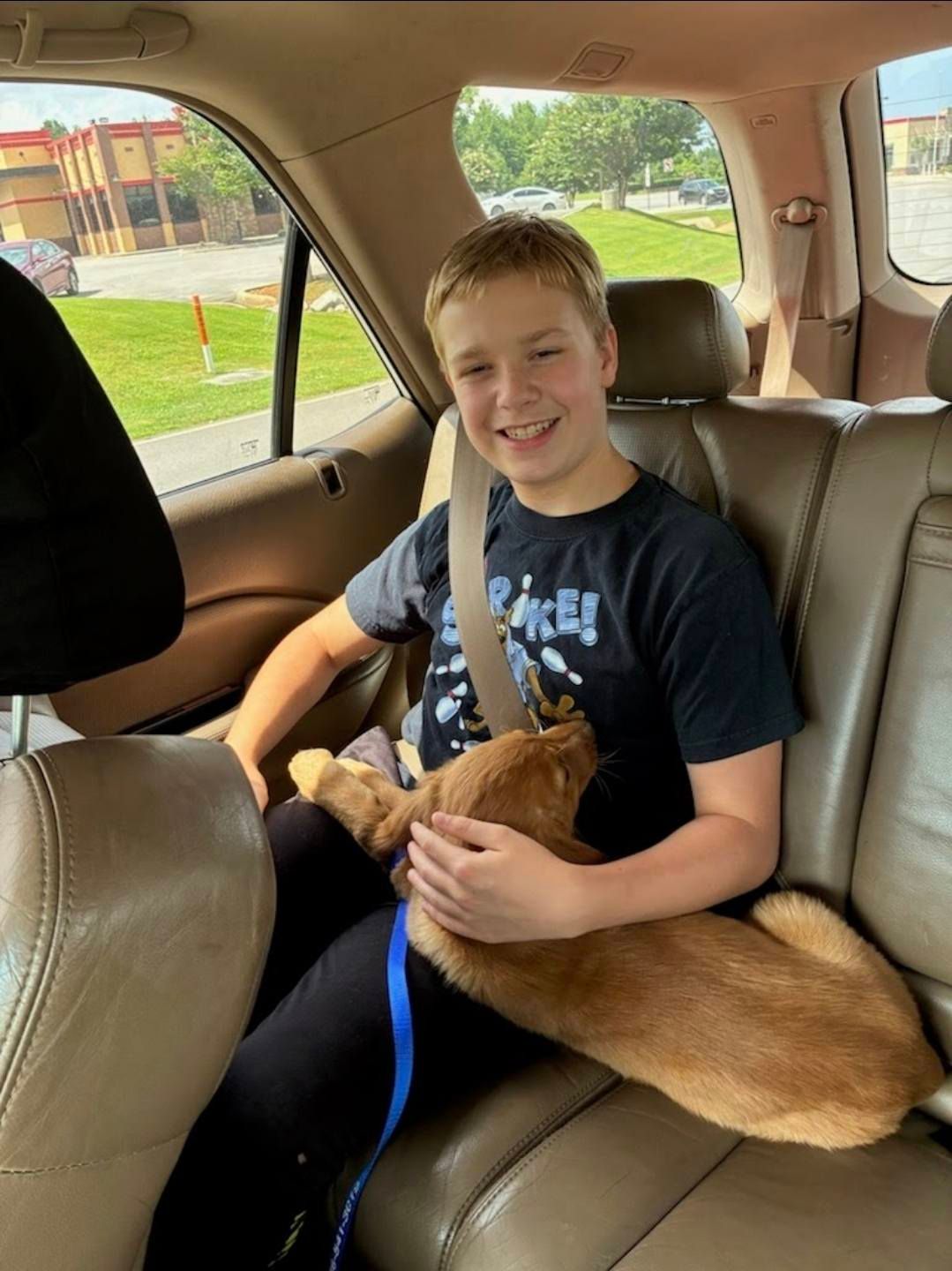 A smiling person sits in the back of a car, holding a light brown dog in their lap.