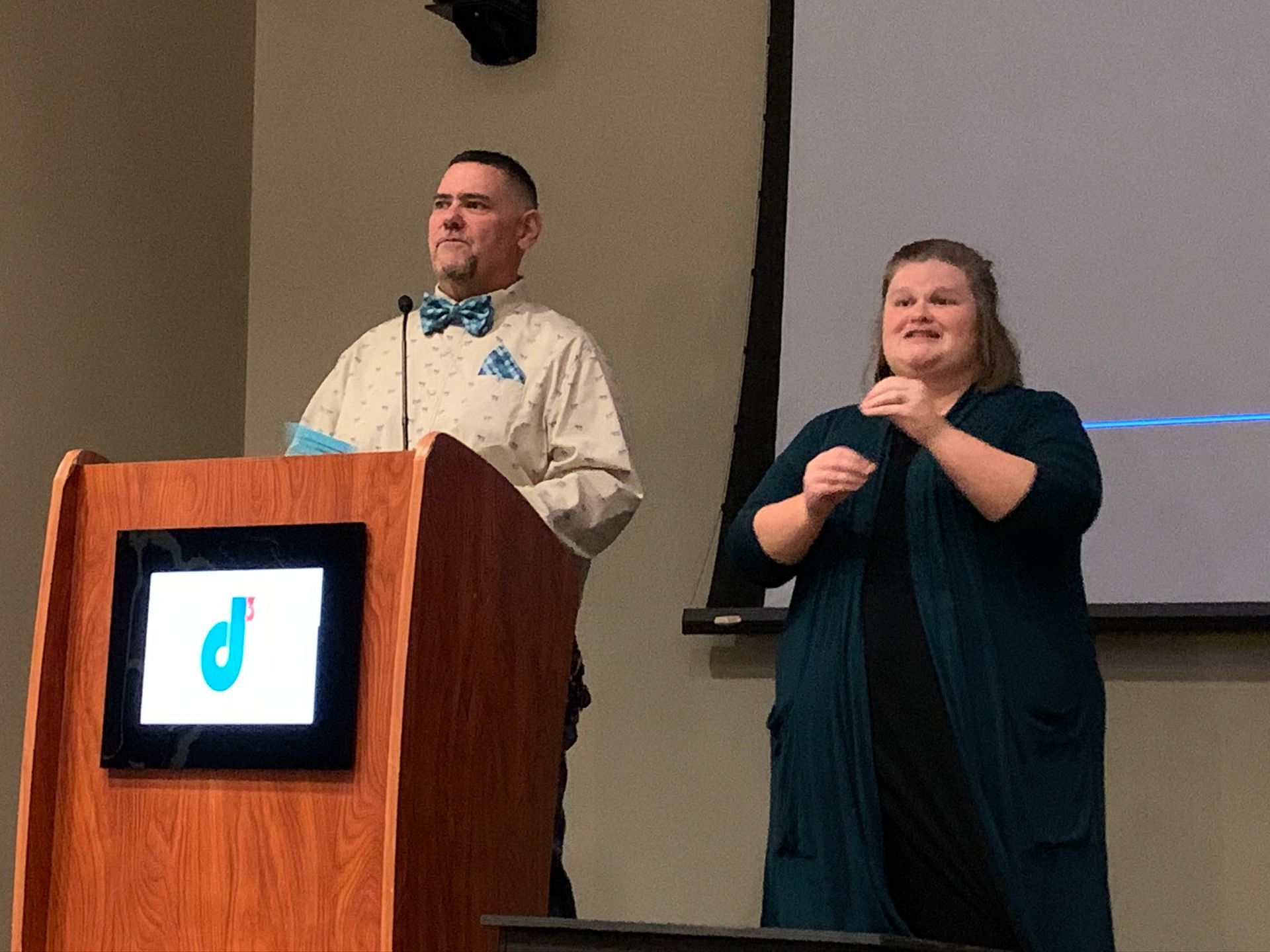 A person in a bow tie speaks at a wooden podium with a screen logo, while someone signs in American Sign Language nearby.