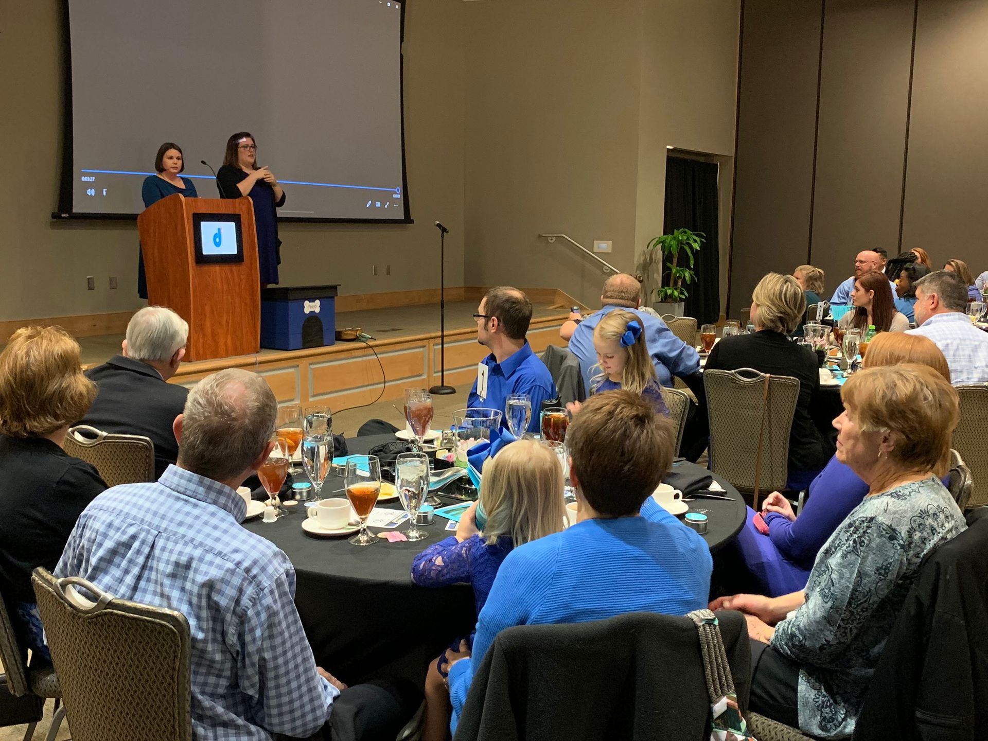Speakers at a podium address an audience seated at round tables in a conference room.