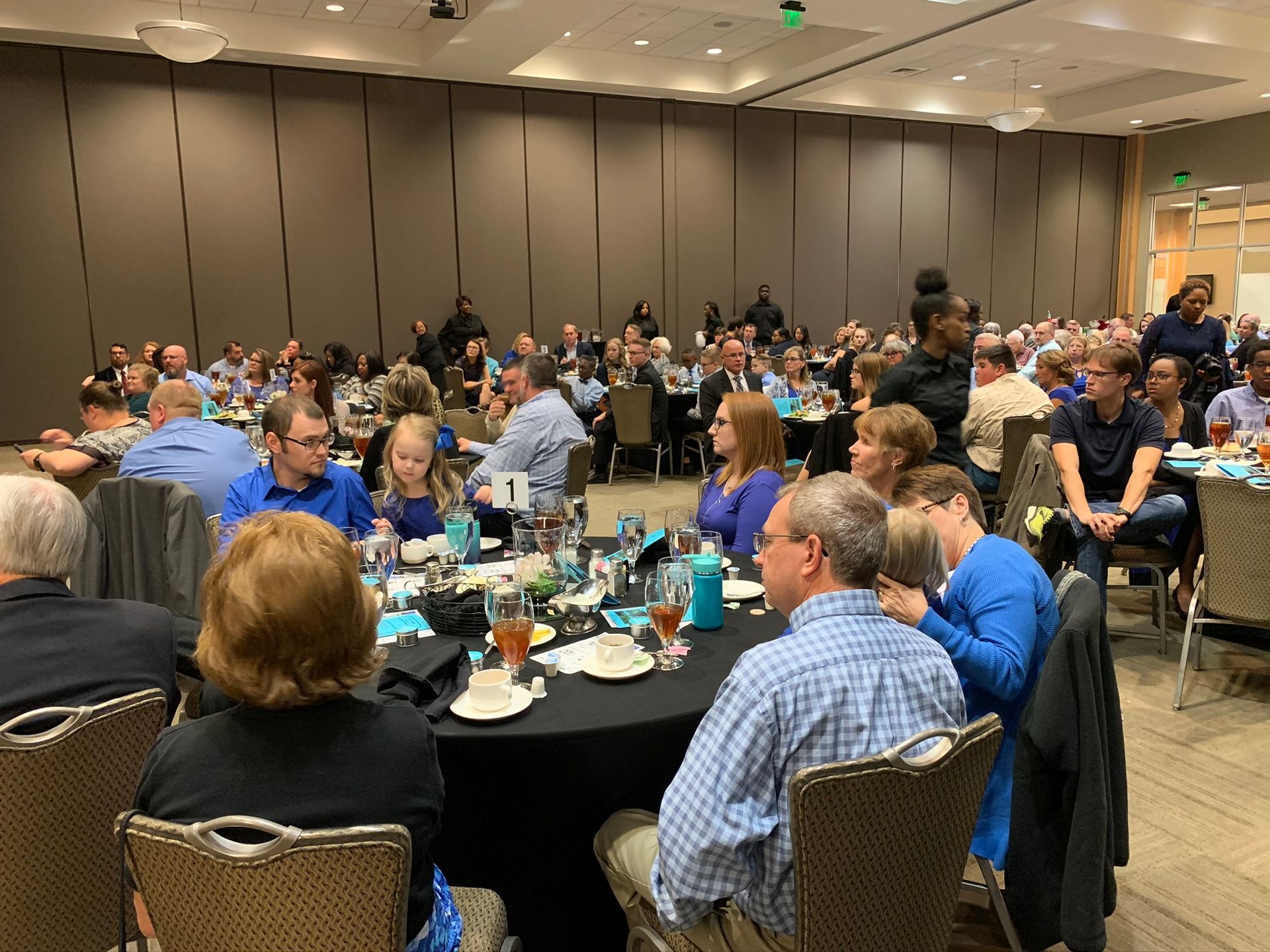 A large group of people sit at round tables in a conference room, listening to a presentation.