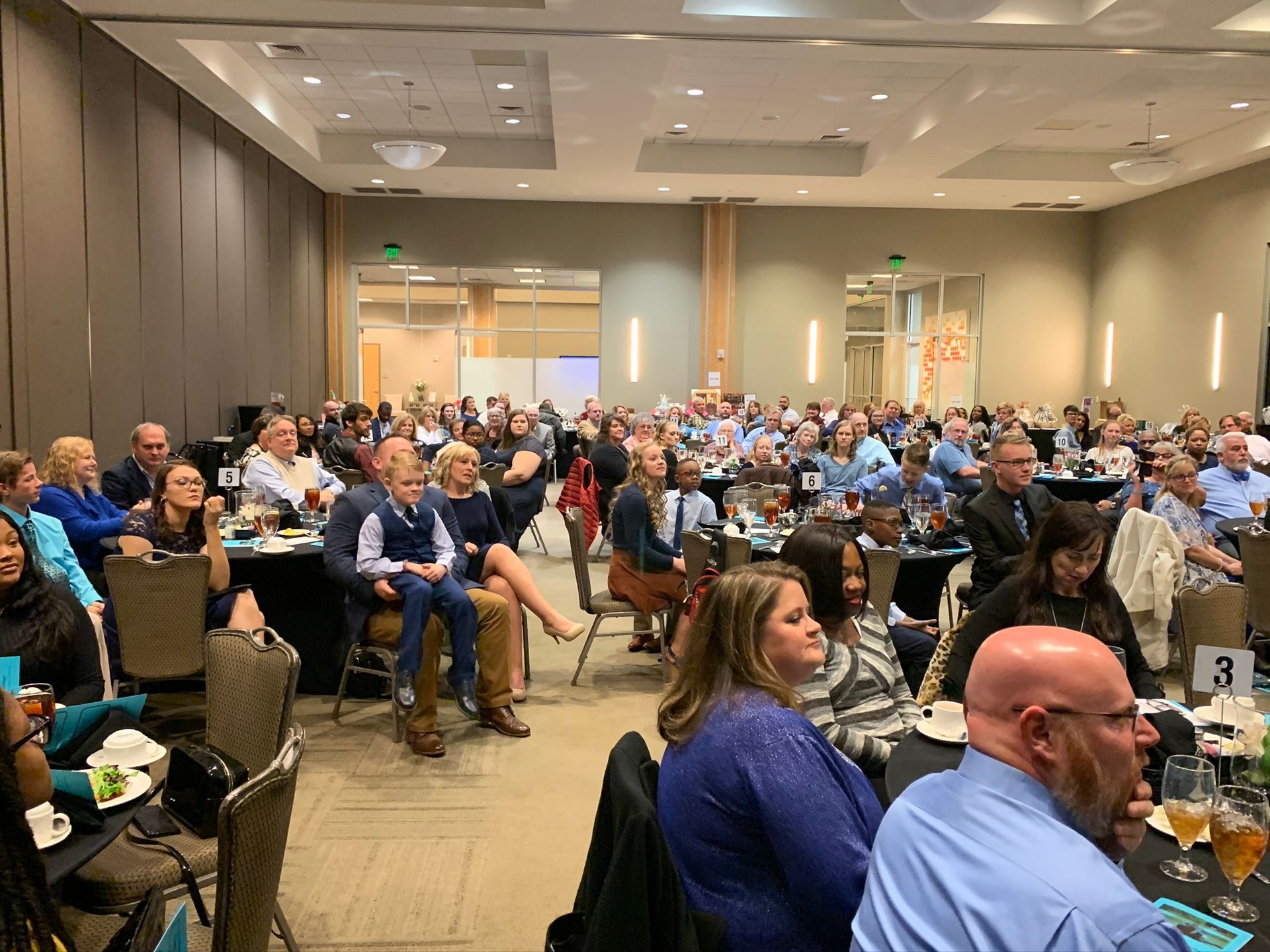 A large audience sits at round tables in a brightly lit banquet hall, watching a presentation from the back of the room.