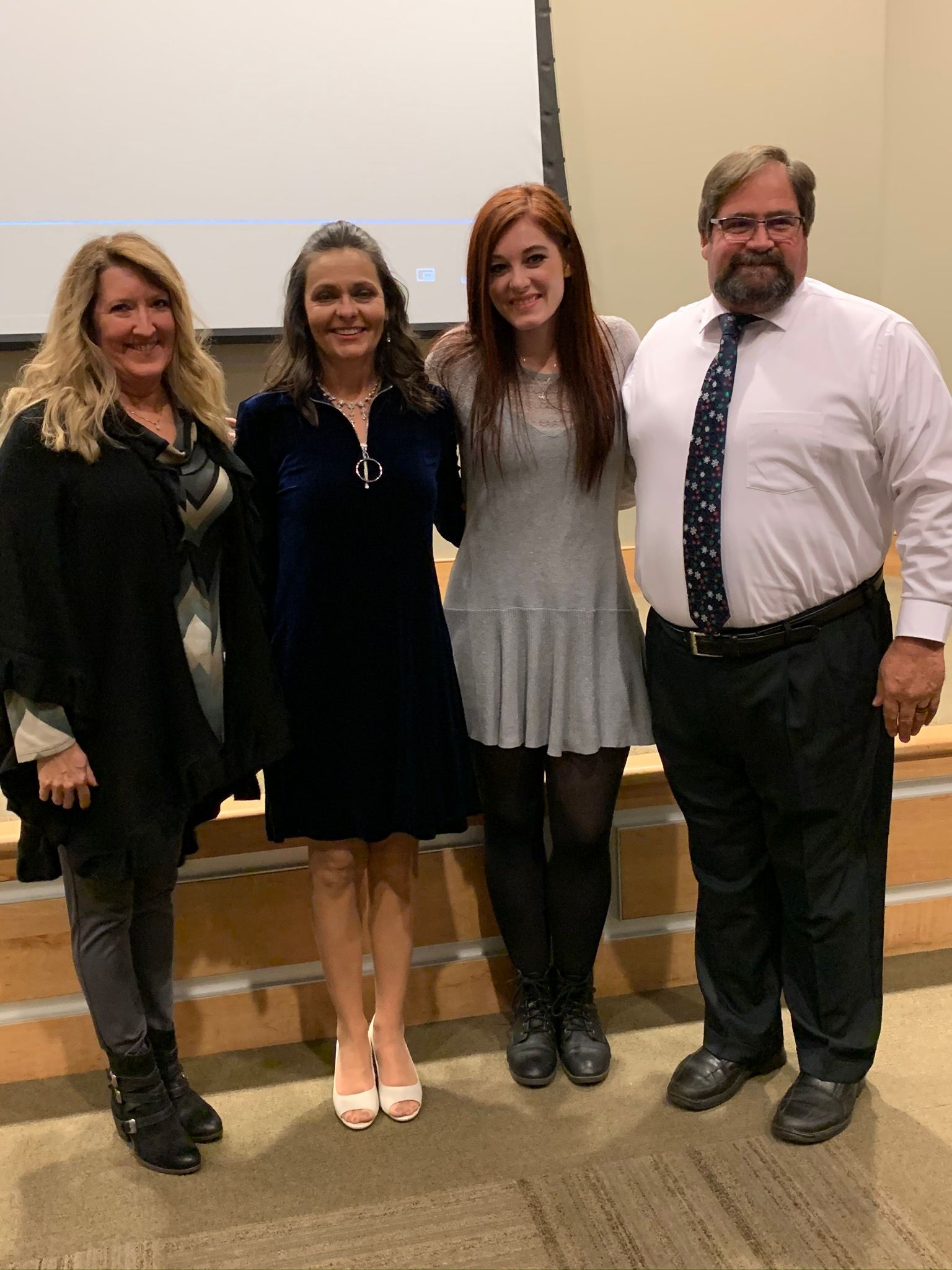 Four people stand in a line against a plain wall, smiling at the camera during an indoor event.