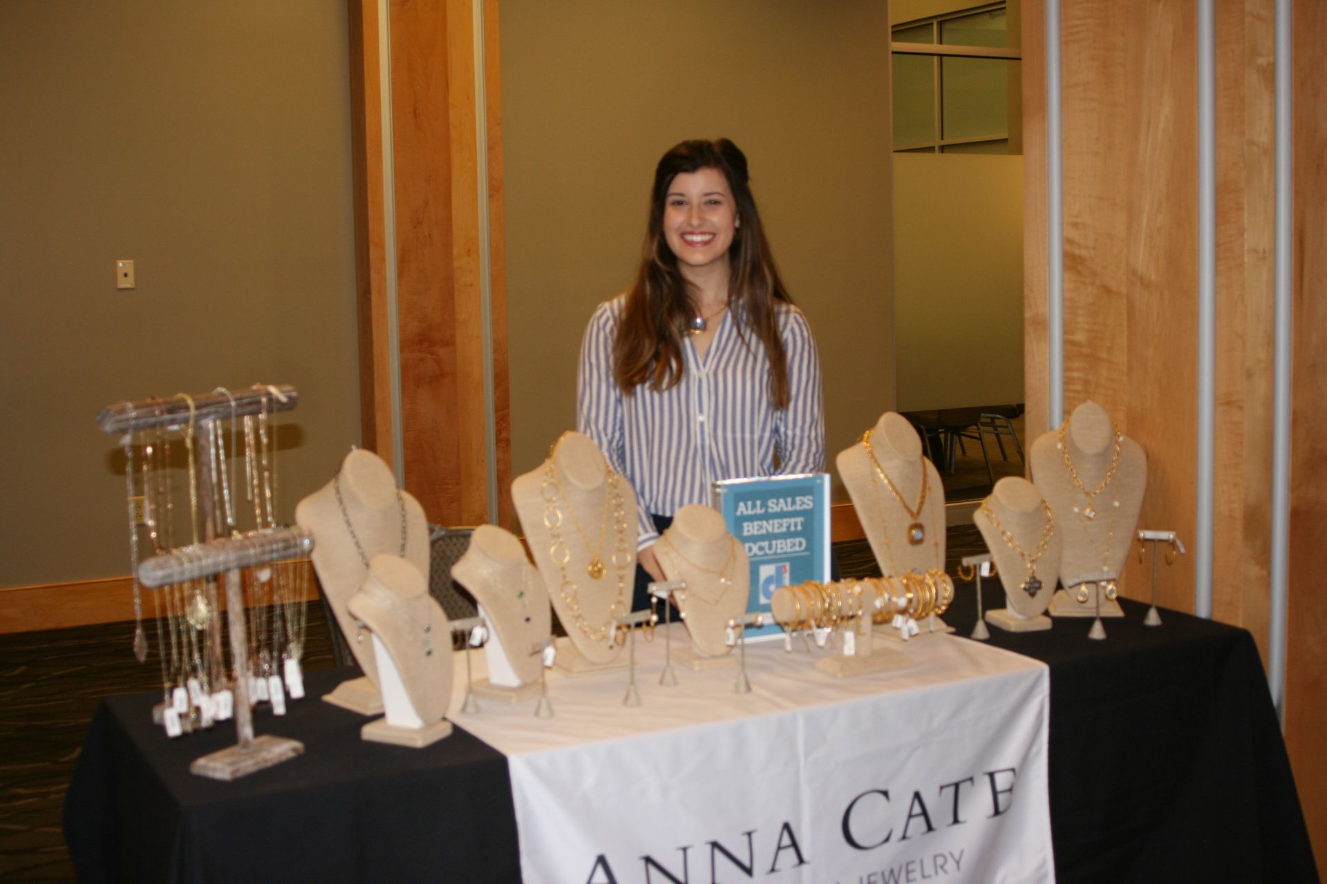 A person smiles behind a table displaying necklaces and jewelry on stands, with an 