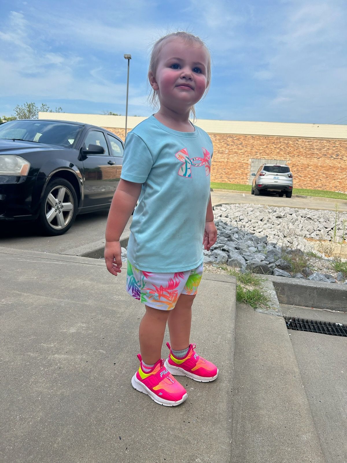 A person in a light blue t-shirt and patterned shorts stands on a sidewalk, wearing bright pink sneakers.