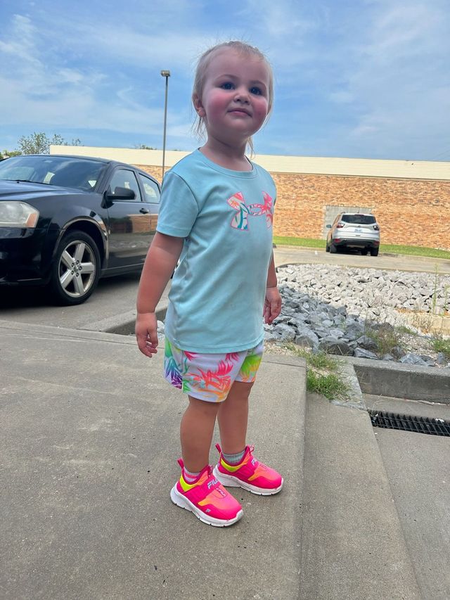 A person in a light blue t-shirt and patterned shorts stands on a sidewalk, wearing bright pink sneakers.