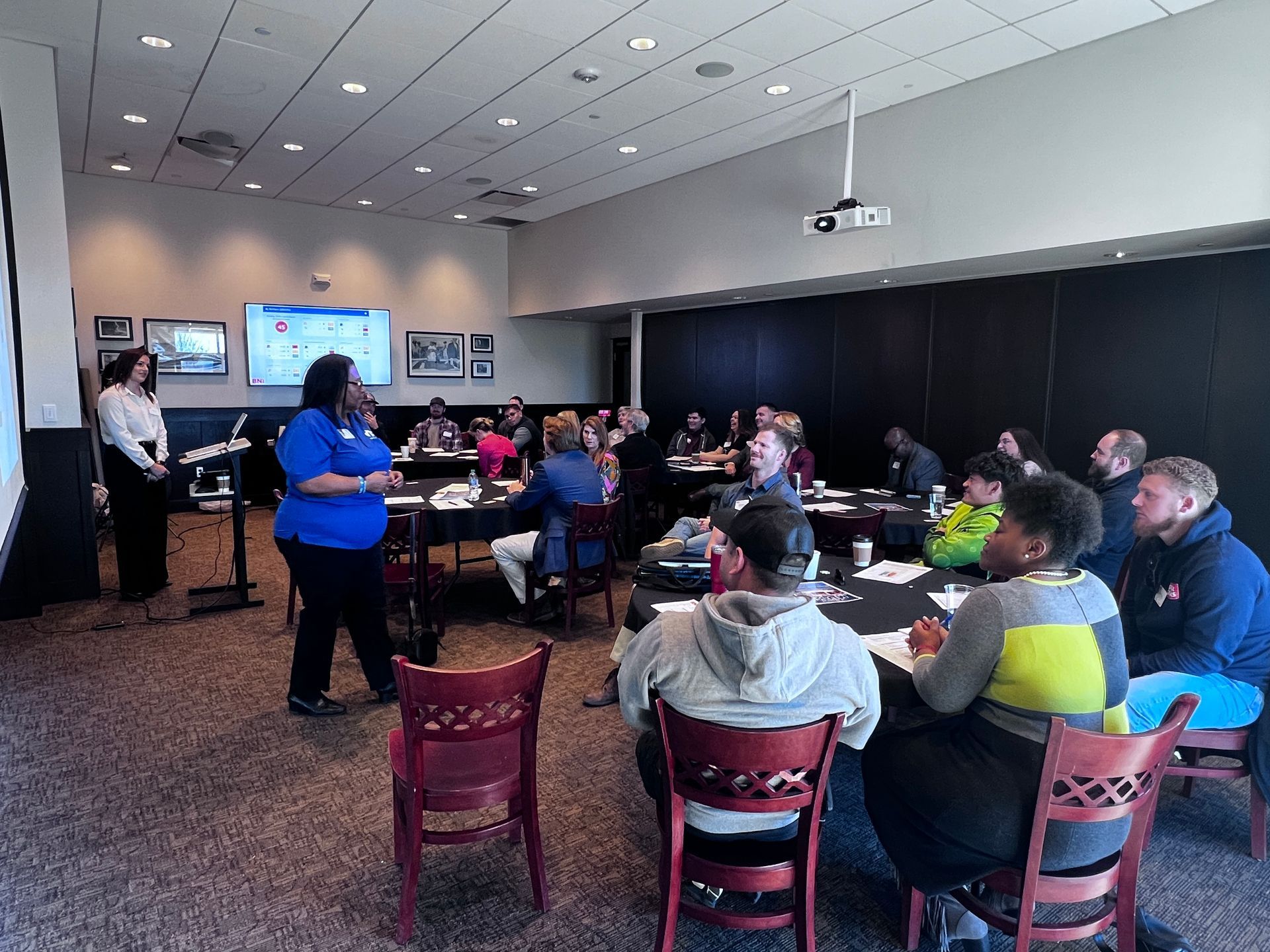 A speaker in a blue shirt addresses a group seated at round tables in a conference room with neutral-toned walls.