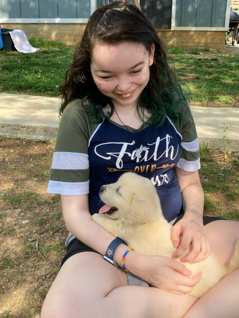 A smiling person sitting on the grass holds a small, light-colored puppy that has its mouth open in a pant.