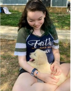 A person sitting cross-legged outdoors holds a happy, light-colored puppy in their lap.
