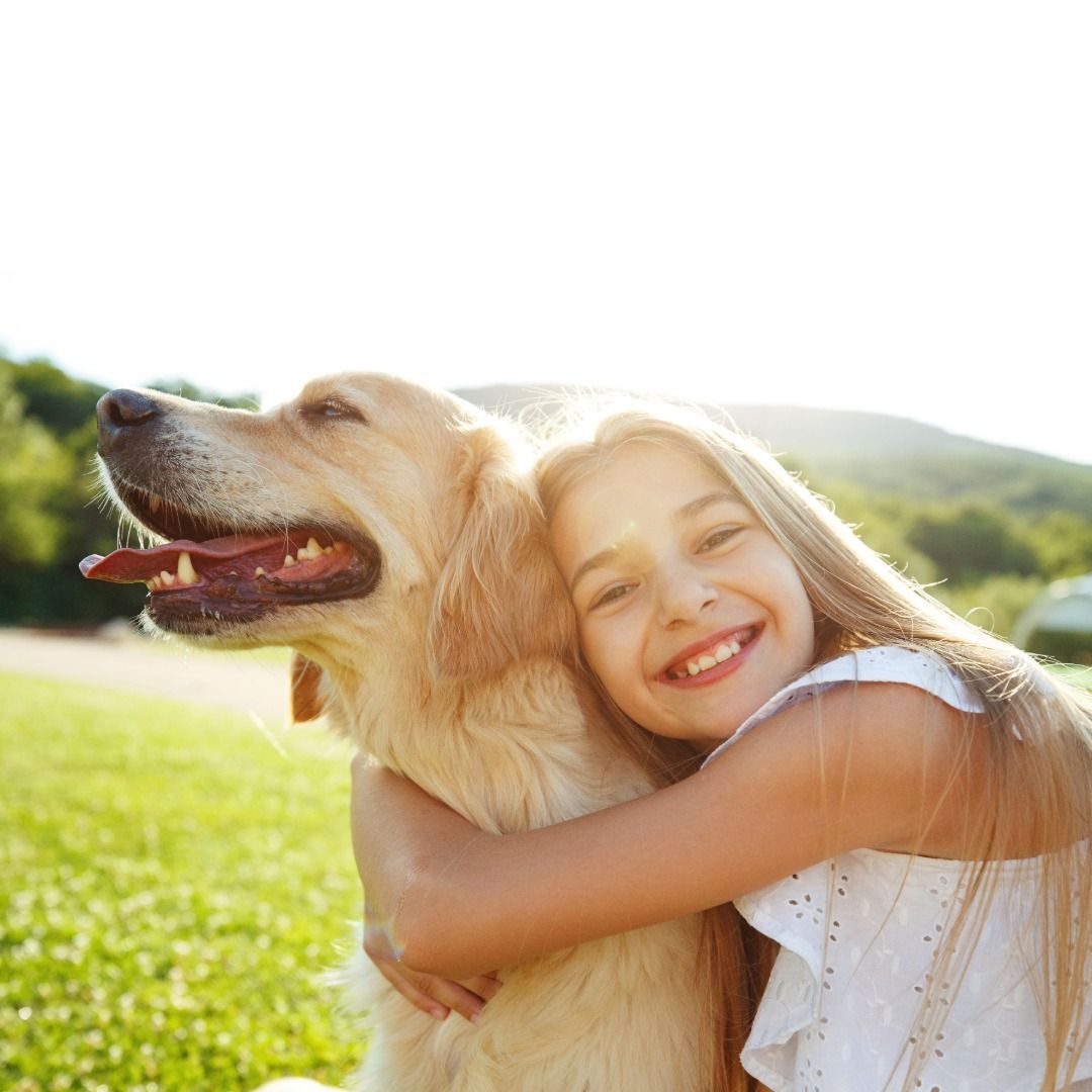 A smiling person with long hair embraces a light-brown dog outdoors in a grassy field under bright sunlight.