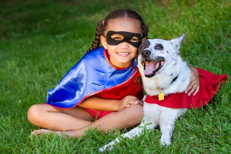 A child in a superhero mask and blue cape sits in the grass hugging a happy, red-caped dog.