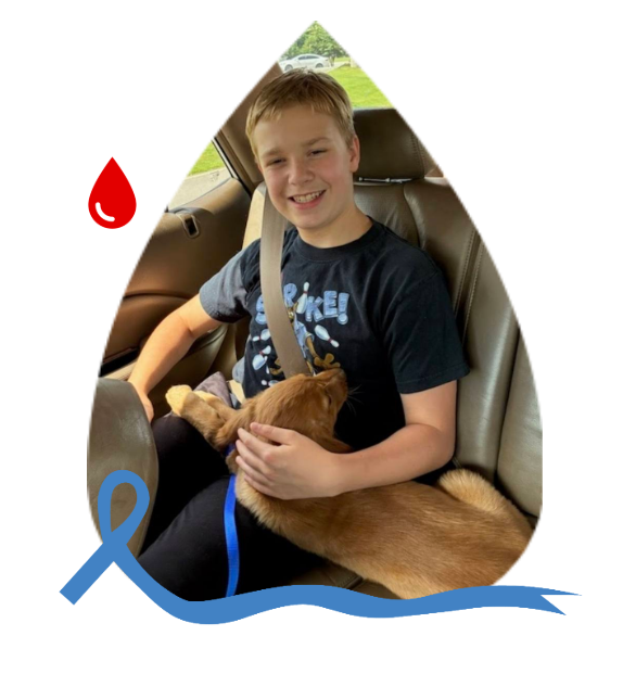 A person sits in a car passenger seat, smiling while holding a puppy. A red blood drop and a blue ribbon frame the image.
