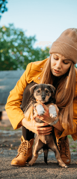 A person in a tan beanie and yellow jacket squats outdoors, gently holding a small, brown and black puppy.