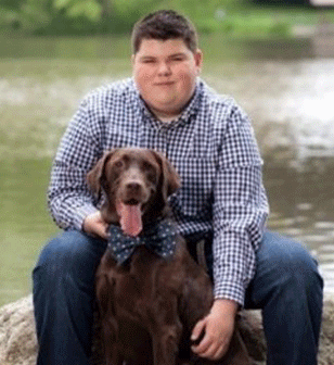 A person wearing a blue plaid shirt and jeans sits outdoors with a brown dog wearing a patterned bow tie.