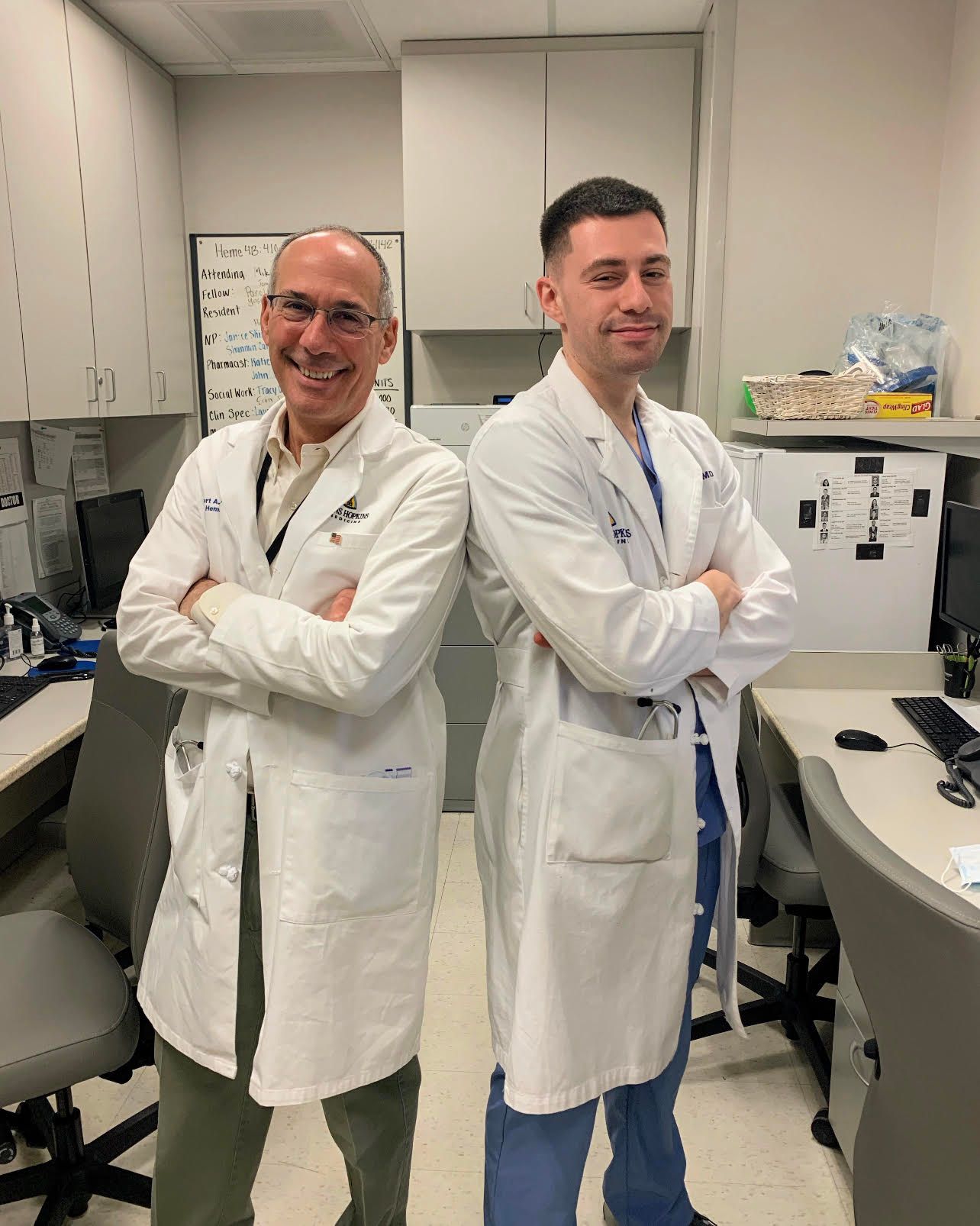 Two doctors are standing next to each other in a room with their arms crossed.