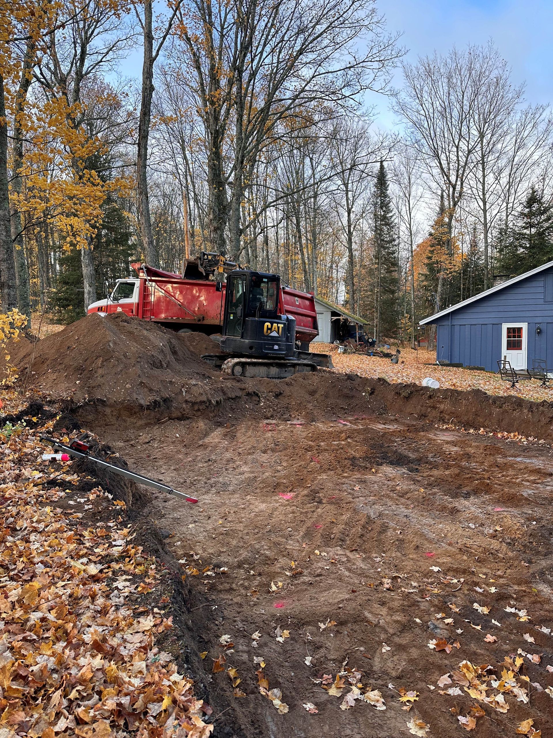 A red dump truck and a black Caterpillar excavator work on a large dirt excavation site next to a blue building.