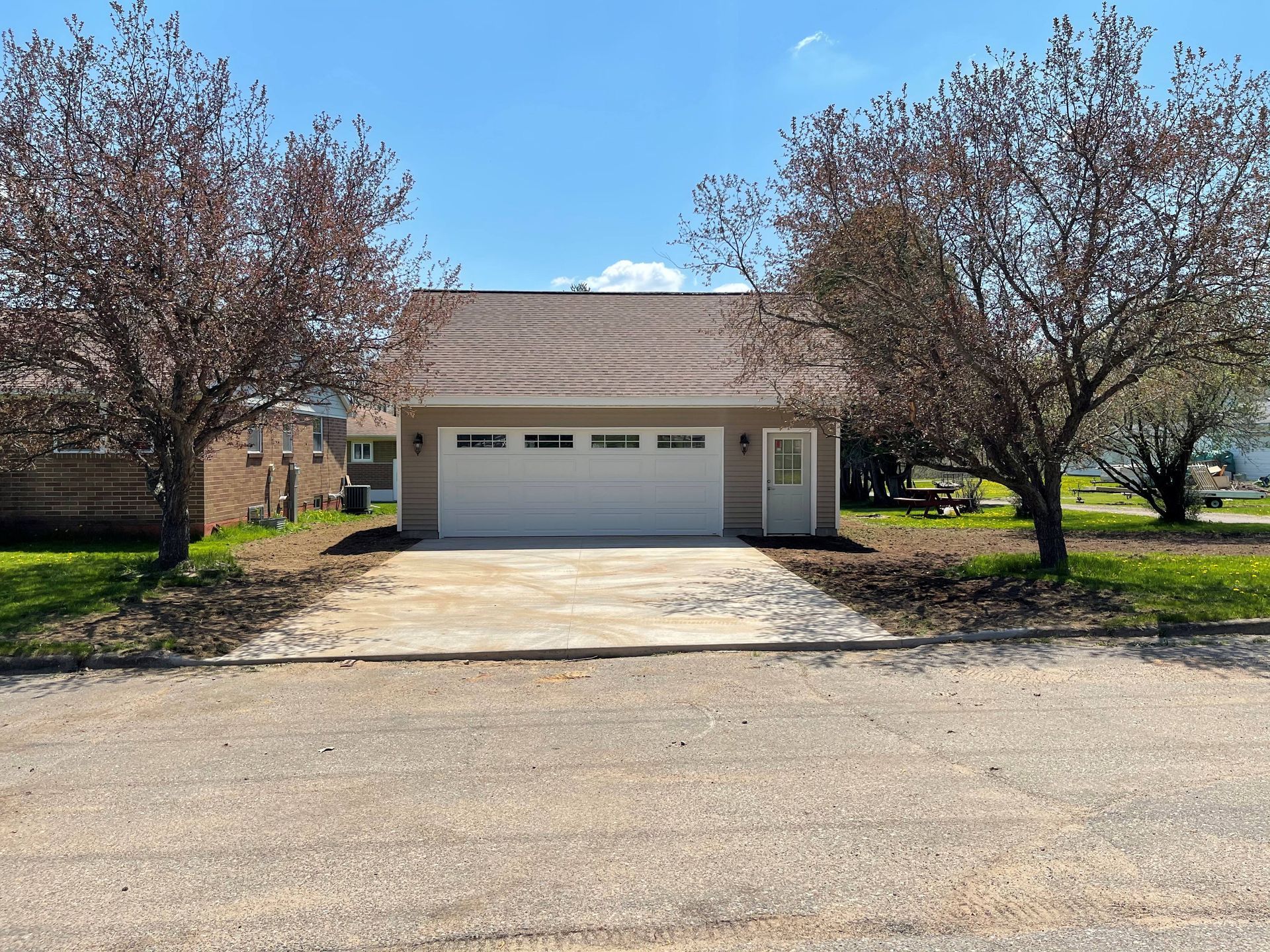 A beige detached garage with a white door and a side entry door, centered between two trees under a clear blue sky. A beige detached garage with a white door and a side entry door, centered between two trees under a clear blue sky.