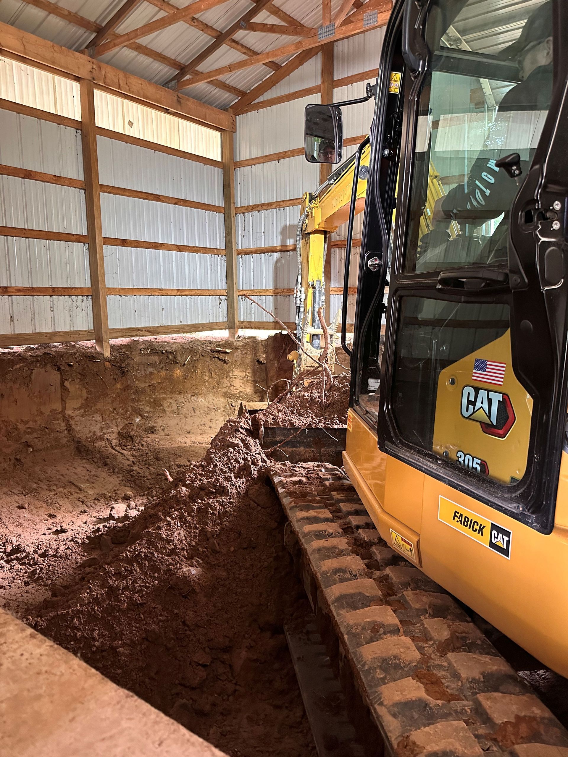 A yellow Caterpillar excavator sits inside a metal-walled barn, digging a large trench in the dirt floor.