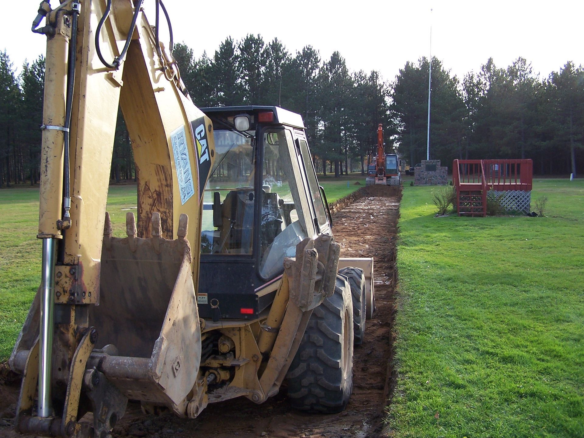 A yellow Caterpillar backhoe parked on a grass field next to a freshly dug trench, with trees and a red deck nearby. A yellow Caterpillar backhoe parked on a grass field next to a freshly dug trench, with trees and a red deck nearby.
