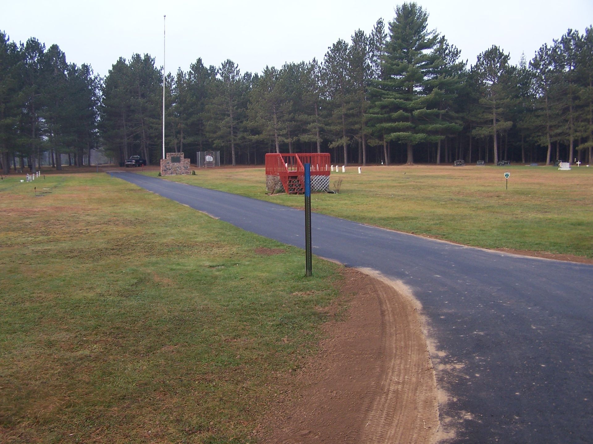 A paved road leads through a grassy cemetery toward a red committal shelter, a flagpole, and a dense line of pine trees. A paved road leads through a grassy cemetery toward a red committal shelter, a flagpole, and a dense line of pine trees.