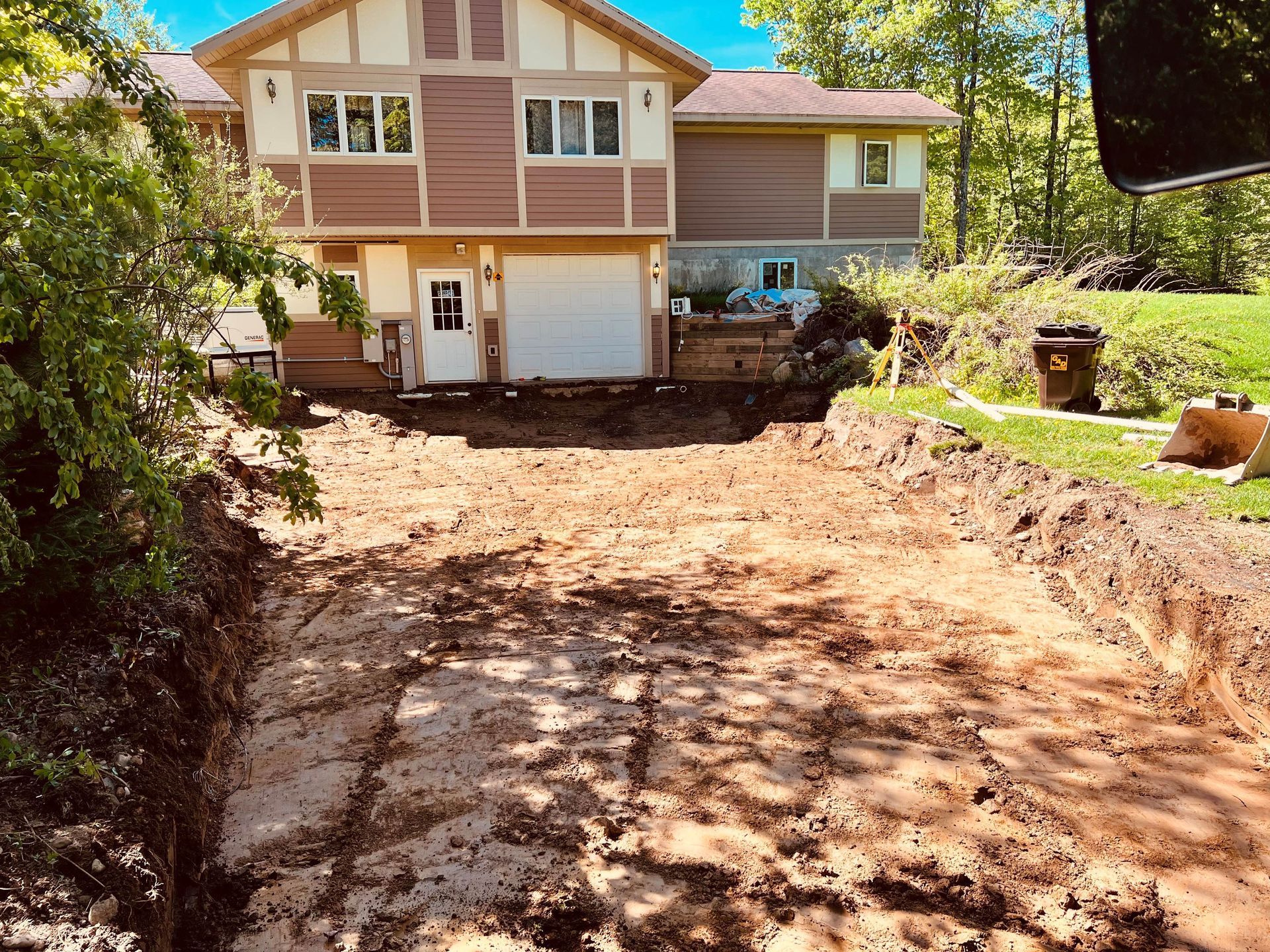 A house under construction with a large, excavated dirt driveway in front, surrounded by trees and greenery. A house under construction with a large, excavated dirt driveway in front, surrounded by trees and greenery.