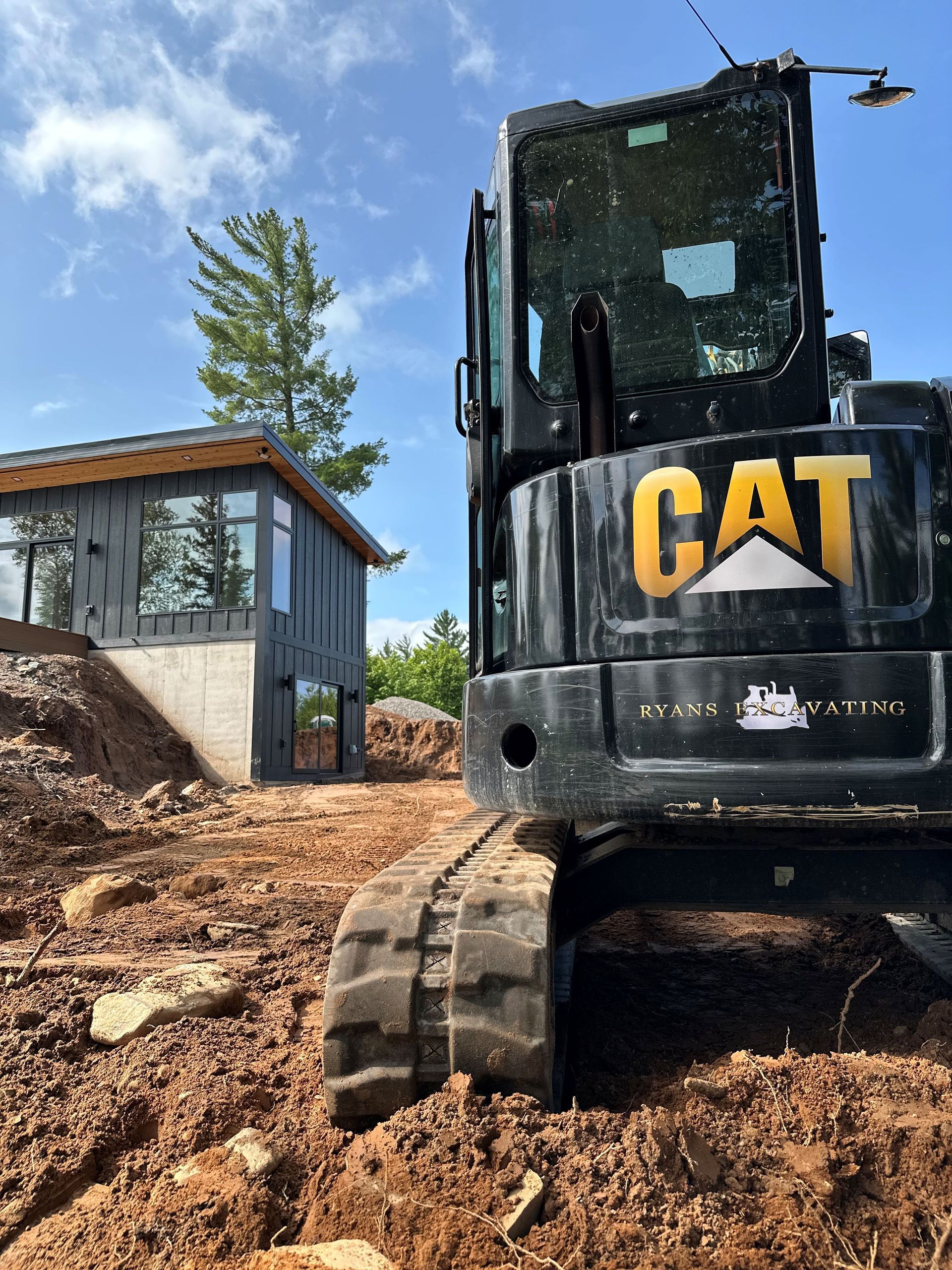 A black CAT excavator parked on a dirt construction site in front of a modern home with large windows.