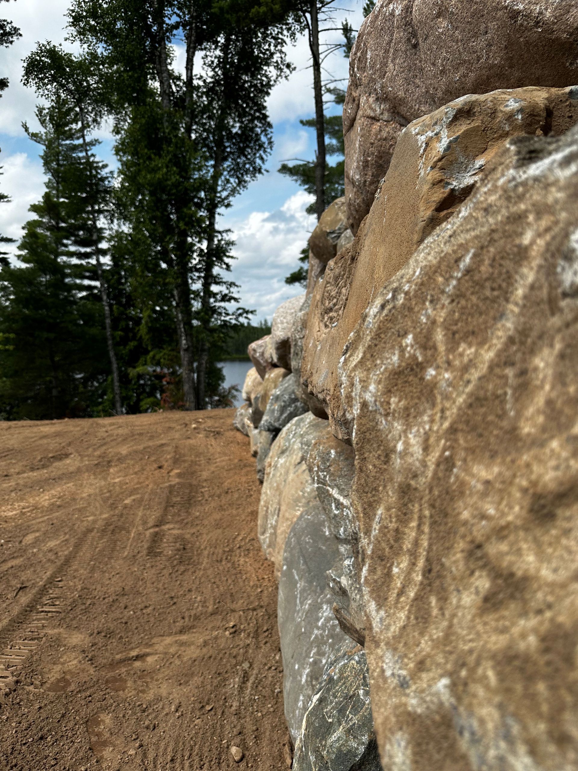 A low-angle view of a stacked stone retaining wall bordering a dirt area, with trees and a lake in the background.