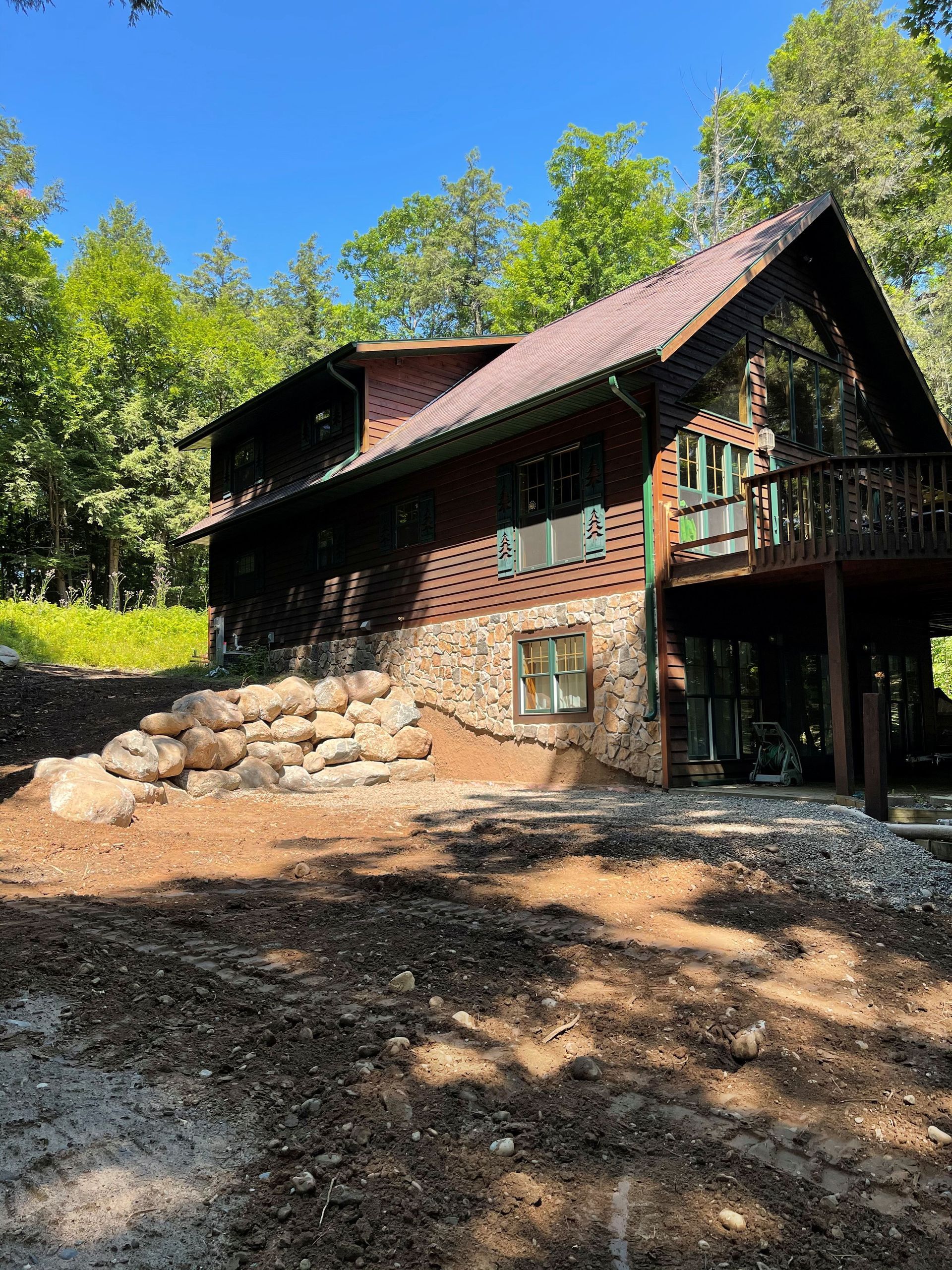 A brown rustic cabin with a stone base, a wooden deck, and a sloping roof, set in a wooded area with a dirt landscape.